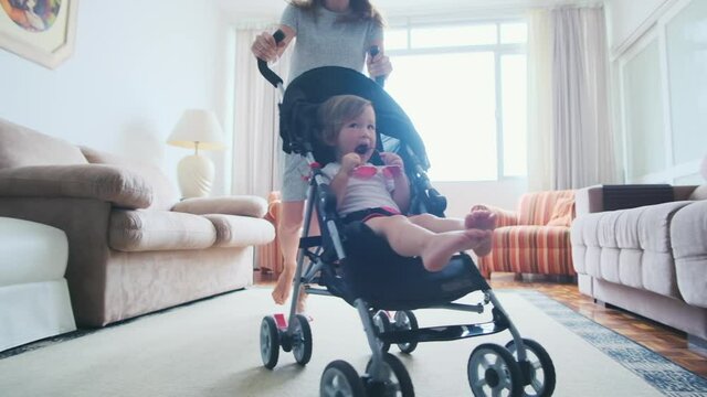 Family have fun in room. Mother gives her toddler girl a ride on the baby stroller along the rooms in apartment