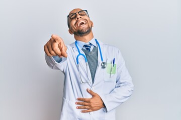 Hispanic adult man wearing doctor uniform and stethoscope laughing at you, pointing finger to the camera with hand over body, shame expression