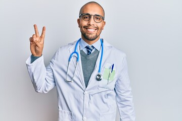 Hispanic adult man wearing doctor uniform and stethoscope smiling looking to the camera showing fingers doing victory sign. number two.