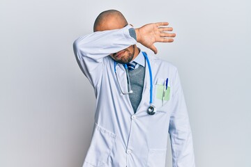 Hispanic adult man wearing doctor uniform and stethoscope covering eyes with arm, looking serious and sad. sightless, hiding and rejection concept