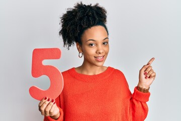 Young african american girl holding number five smiling happy pointing with hand and finger to the side - Powered by Adobe