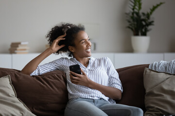 Smiling young African American woman sit relax on couch at home use modern smartphone gadget. Happy millennial biracial female have fun browsing internet on cellphone rest in living room on weekend.