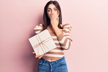Young beautiful girl holding gift covering mouth with hand, shocked and afraid for mistake....