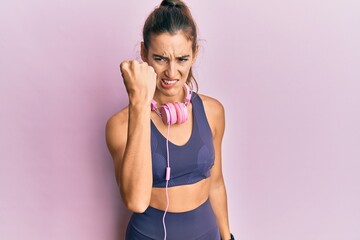 Young beautiful woman wearing gym clothes and using headphones angry and mad raising fist frustrated and furious while shouting with anger. rage and aggressive concept.