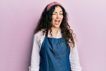 Young brunette woman with curly hair wearing cleaner apron and gloves winking looking at the camera...