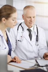 Fototapeta premium Doctors colleagues and elderly therapist discussing current disease therapy while sitting at working place in sunny clinic. Teamwork in medicine