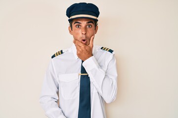 Young hispanic man wearing airplane pilot uniform looking fascinated with disbelief, surprise and amazed expression with hands on chin