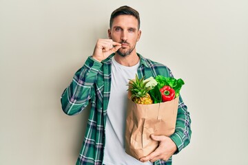 Handsome man with beard holding paper bag with groceries mouth and lips shut as zip with fingers....
