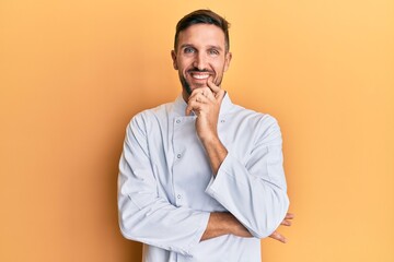 Handsome man with beard wearing professional cook uniform looking confident at the camera with smile with crossed arms and hand raised on chin. thinking positive.