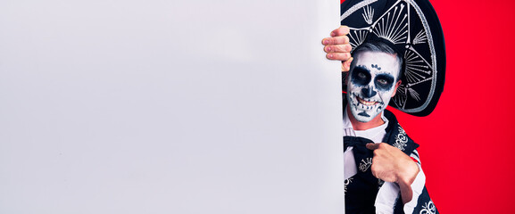 Young man wearing mexican day of the dead costume holding blank empty banner pointing finger to one self smiling happy and proud
