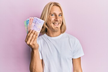 Caucasian young man with long hair holding thai baht banknotes looking positive and happy standing and smiling with a confident smile showing teeth