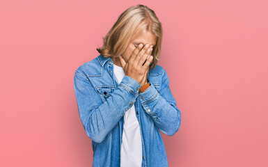 Caucasian man with blond long hair wearing casual denim jacket with sad expression covering face with hands while crying. depression concept.