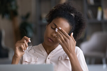 Exhausted young African American female employee massage eyes suffer from headache or migraine...