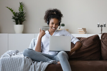 Smiling African American woman in earphones sit on sofa at home talk on video call on laptop. Happy...
