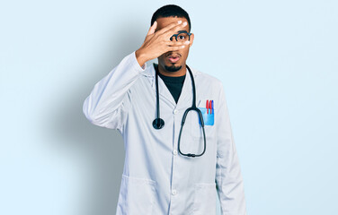 Fototapeta premium Young african american man wearing doctor uniform and stethoscope peeking in shock covering face and eyes with hand, looking through fingers with embarrassed expression.