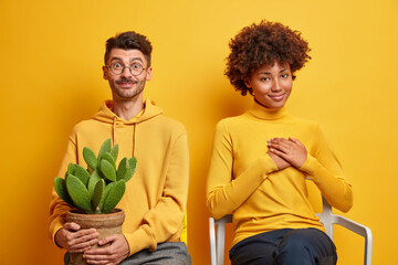 Isolated shot of mixed race woman and man sit next to each other on comfortable chairs isolated over yellow background. Cheerful guy wears sweatshirt holds potted cactus female makes thankful gesture