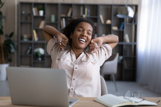 Happy young African American woman stretch relax in chair at home office workplace finish job on computer on time. Smiling biracial female rest from laptop job, take break breathe fresh air.