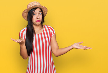 Young hispanic woman wearing summer hat clueless and confused expression with arms and hands...