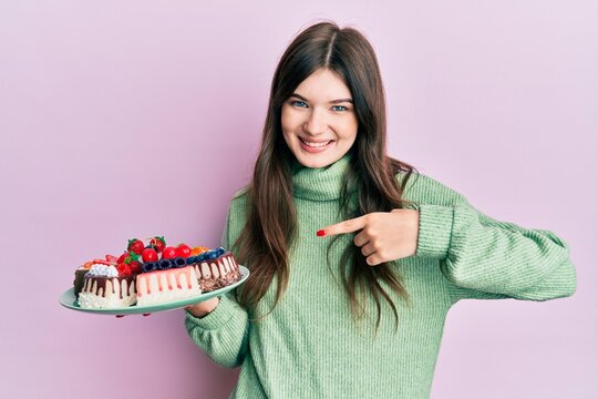 Young beautiful caucasian girl holding cake slices pointing finger to one self smiling happy and proud