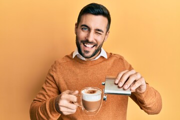 Young hispanic man drinking a cup of coffee with alcohol winking looking at the camera with sexy...
