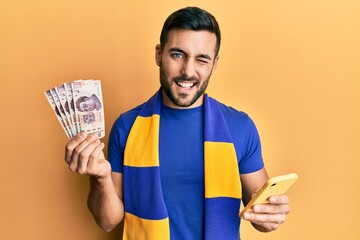 Young hispanic man football supporter using smartphone holding mexican pesos banknotes winking...