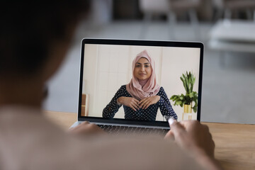 Back view of female employee talk speak on video call on laptop with Muslim Indian colleague. Woman...