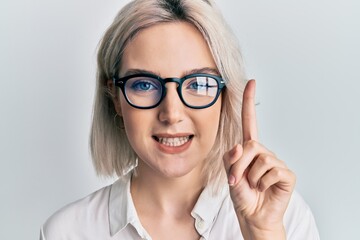 Young blonde girl wearing casual clothes and glasses smiling with an idea or question pointing finger with happy face, number one