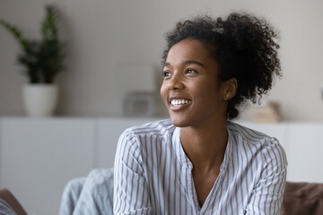 Smiling millennial African American woman relax in cozy own living room look in distance dreaming or visualizing. Happy young biracial female renter think or plan. Vision, visualization concept. - Powered by Adobe