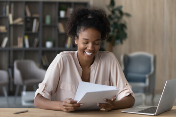 Happy young African American female sit at desk at home office read consider paper documents or...