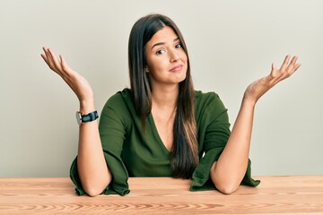 Young brunette woman wearing casual clothes sitting on the table clueless and confused expression...