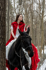 beautiful girl in a white dress and a red cloak with a black horse in the spring forest