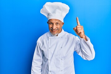 Middle age grey-haired man wearing professional cook uniform and hat pointing finger up with successful idea. exited and happy. number one.