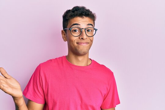Young handsome african american man wearing glasses over pink background clueless and confused expression with arms and hands raised. doubt concept.