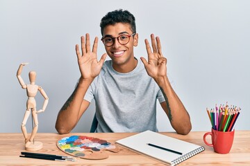 Young handsome african american man painter sitting palette and art manikin showing and pointing up with fingers number nine while smiling confident and happy.