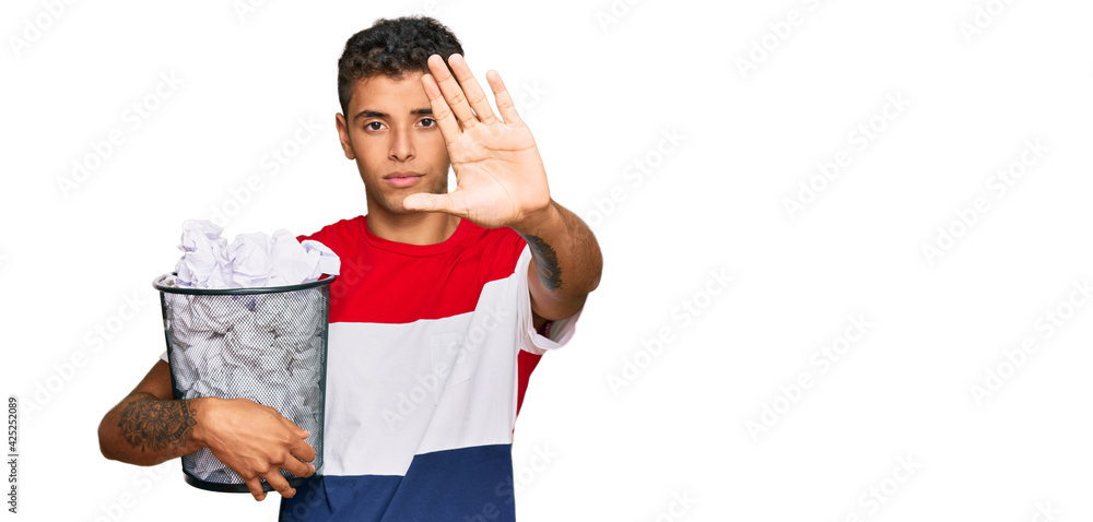 Sticker Young handsome african american man holding paper bin full of crumpled papers with open hand doing stop sign with serious and confident expression, defense gesture