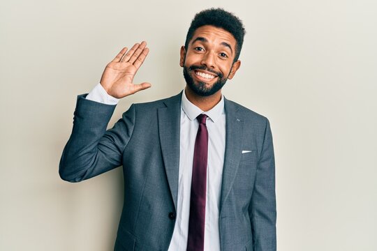 Handsome hispanic man with beard wearing business suit and tie waiving saying hello happy and smiling, friendly welcome gesture