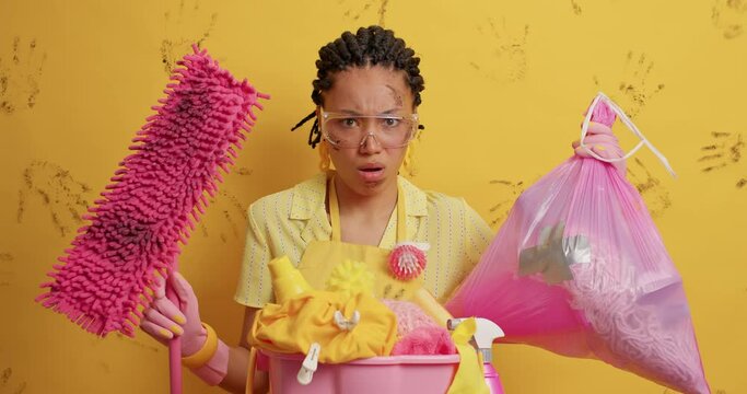 Displeased tired dark skinned lady hates dirt looks at dirty mop and garbage bag wears transparent glasses smirks face from aversion isolated over yellow background. Puzzled ethnic housewife
