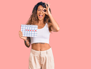 Young hispanic woman with tattoo holding heart calendar smiling happy doing ok sign with hand on eye looking through fingers