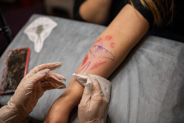 Master tattoo draws the red paint on the clients tattoo. Tattoo artist holding a pink tattoo machine in black sterile gloves and working on the professional blue mat.