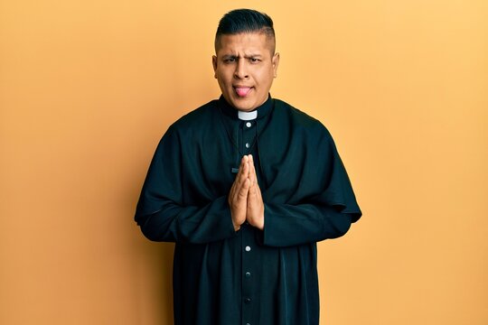 Young latin priest man praying with hands together sticking tongue out happy with funny expression.
