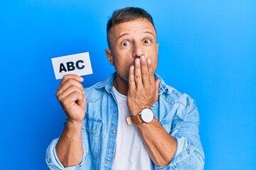 Handsome muscle man holding abc letters on paper covering mouth with hand, shocked and afraid for mistake. surprised expression