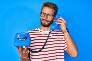 Handsome caucasian man with beard holding vintage telephone in shock face, looking skeptical and...