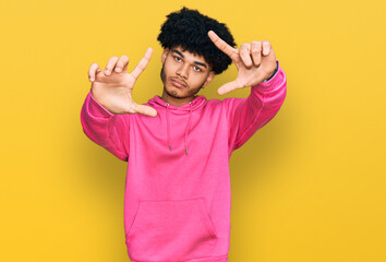 Young african american man with afro hair wearing casual pink sweatshirt doing frame using hands palms and fingers, camera perspective