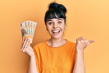 Young hispanic girl holding norwegian krone banknotes pointing thumb up to the side smiling happy...