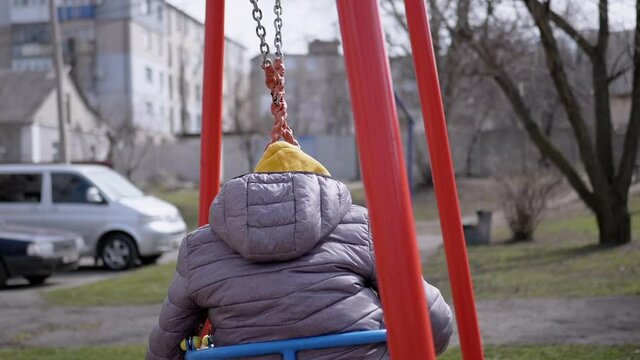Happy, Laughing Child Swinging on a Swing in the Yard Without Friends. Boy in a gray jacket and cap. Children are sitting at home playing video games. Sunny day. Childhood in quarantine. Slow-motion.