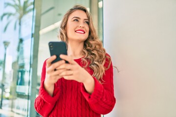 Young blonde girl smiling happy using smartphone standing at the city.