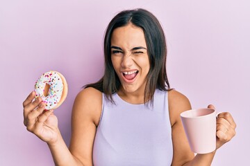 Young brunette woman eating doughnut and drinking coffee winking looking at the camera with sexy...