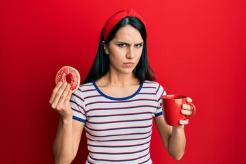 Young hispanic woman eating doughnut and drinking coffee skeptic and nervous, frowning upset...