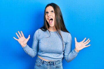 Young brunette teenager wearing casual sweater crazy and mad shouting and yelling with aggressive expression and arms raised. frustration concept.