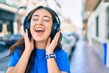 Young hispanic woman smiling happy listening to music using headphones walking at the city.